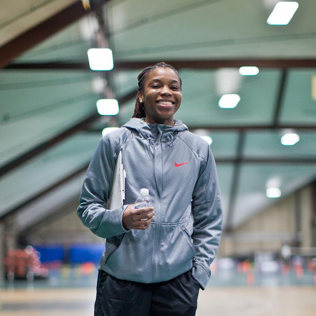 Priscilla Edwards is standing on a basketball court, holding a bottle of water and a clip board, she is smiling directly at the camera.